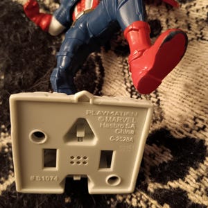 May include: A gray plastic base with the text "Playmation", "&copy;MARVEL", "Hasbro SA", "China", "C-2528A", and "#B1074". A red, white, and blue Captain America action figure stands on the base.
