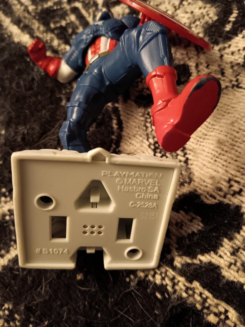 May include: A gray plastic base with the text "Playmation", "&copy;MARVEL", "Hasbro SA", "China", "C-2528A", and "#B1074". A red, white, and blue Captain America action figure stands on the base.