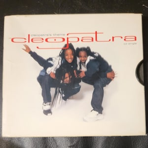 May include: CD single for "Cleopatra's Theme" featuring three people. The cover has a white background with the band's name in red. The people are wearing dark jackets and jeans. The CD case is white.