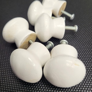 May include: A collection of white, oval-shaped wooden cabinet knobs with attached silver screws. The knobs are painted with a glossy white finish and are arranged on a dark, textured surface. These knobs are suitable for furniture restoration or new builds.