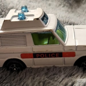 Puede incluir: Coche de policía de juguete blanco con un letrero de POLICÍA rojo y blanco en el lateral. El coche tiene una luz azul claro intermitente en la parte superior.