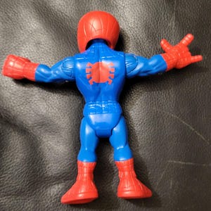May include: A blue and red Spider-Man action figure, viewed from the back. The figure has a red helmet and gloves, with a spider emblem on the back. The legs and arms are outstretched.