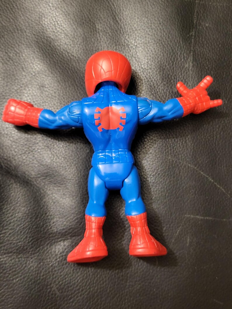 May include: A blue and red Spider-Man action figure, viewed from the back. The figure has a red helmet and gloves, with a spider emblem on the back. The legs and arms are outstretched.