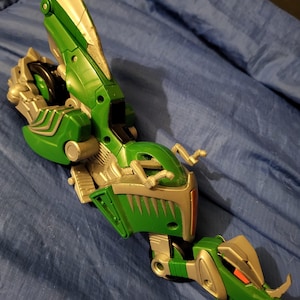 May include: A green and silver toy dragon with articulated sections. The dragon has a detailed design with a head, body, and tail. The toy is made of plastic and has red accents on the head.