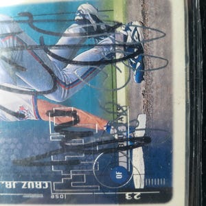 Puede incluir: Una tarjeta de béisbol con un jugador en uniforme azul y blanco, con el nombre "CRUZ JR." visible. La tarjeta está en una funda protectora. El fondo muestra un campo de béisbol.