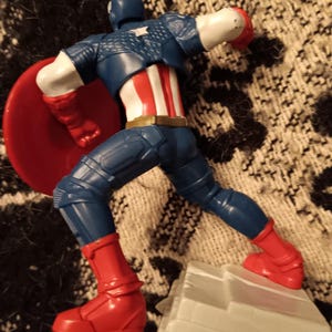 May include: A plastic action figure of Captain America in a blue and red suit, holding a red, white, and blue shield. The figure is standing on a gray base.