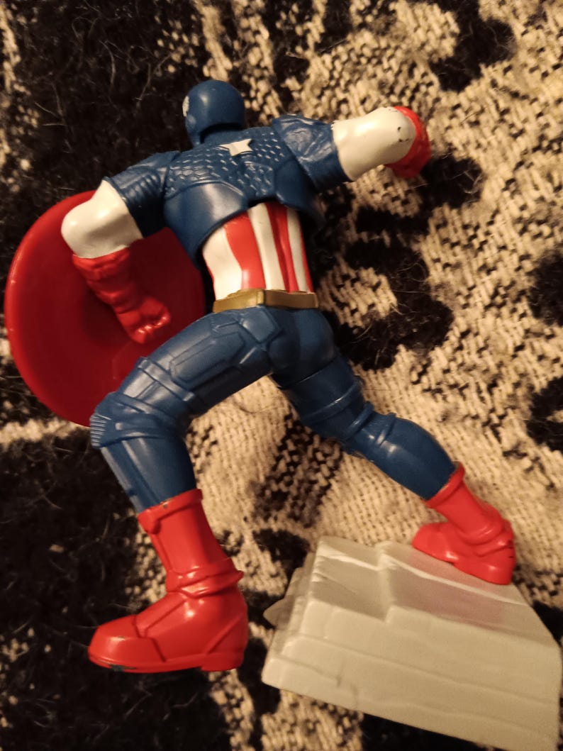 May include: A plastic action figure of Captain America in a blue and red suit, holding a red, white, and blue shield. The figure is standing on a gray base.