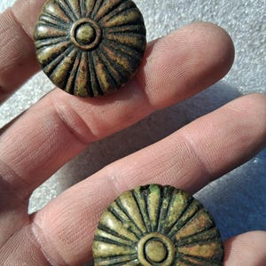 May include: Two circular, bronze-toned metal buttons featuring a raised, radiating sunburst or floral petal pattern. Each button has a smooth, raised central circular disc surrounded by deep, textured grooves extending to the outer edge.