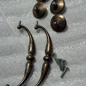 May include: Two curved bronze-toned metal drawer pulls with a smooth finish, accompanied by four matching circular mounting plates and four silver-colored metal screws. The hardware features a tapered design with a central bulbous detail.