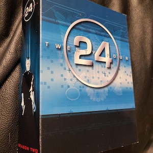 May include: A DVD box set for the television show "24." The box is primarily blue and black, with the show's logo in a white circle. The words "Twenty Four" are printed on the front. The side of the box reads "Season Two."
