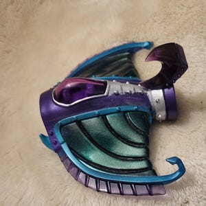 May include: A futuristic helmet in shades of purple and teal, with a curved visor and a claw-shaped protrusion. The helmet has a metallic finish with riveted details and a transparent purple dome. Teal and silver accents enhance the design.