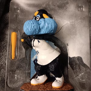 May include: A blue and black baseball mascot figurine holding a bat. The mascot is wearing a white jersey with "Tampa Bay Rays" written on it. The figurine is on a blue and black base.