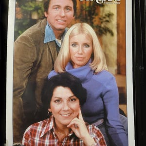May include: A poster for the television show "Three's Company." The image shows three people, with the show's title and "Season One" text visible. The people are smiling. The poster is in a black frame.