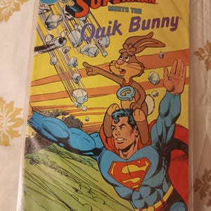 Può includere: Copertina di fumetto con Superman e il Quik Bunny. Superman, nel suo costume blu e rosso, porta il coniglio. La copertina ha uno sfondo giallo con rocce che cadono e il testo "Superman Meets the Quik Bunny."