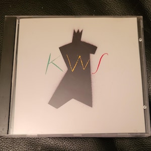May include: A white CD case with a black silhouette of a figure wearing a crown. The figure is outlined in yellow, green, and red. The text "KWS" is written in green, yellow, and red.