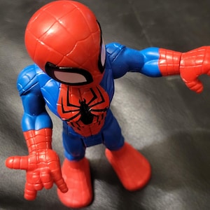 May include: A Spider-Man action figure in a dynamic pose. The figure features a red head and hands, blue torso and legs, and a black spider emblem on the chest. The figure is made of plastic and is a collectible toy.