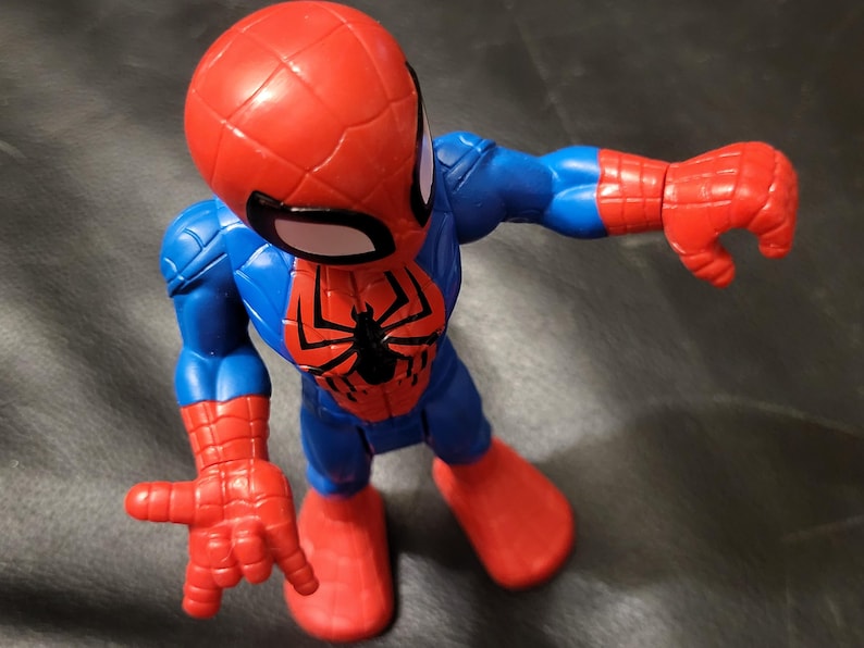 May include: A Spider-Man action figure in a dynamic pose. The figure features a red head and hands, blue torso and legs, and a black spider emblem on the chest. The figure is made of plastic and is a collectible toy.