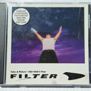 May include: A CD case featuring the album art for "Take A Picture" by Filter. The image shows a person with arms raised against a purple and starry background. The text includes "Filter" and "Includes four exclusive postcards."