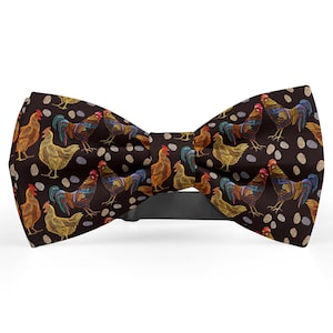 Può includere: Un papillon marrone scuro con un motivo ripetuto di galli e galline colorati, insieme a forme di uova bianche e beige. Il papillon ha un cinturino nero regolabile.