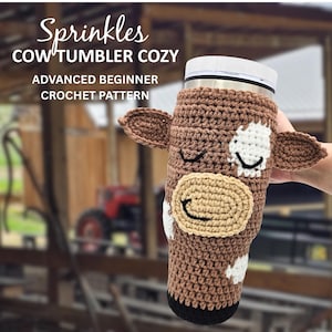 May include: A brown and white crochet cow tumbler cozy. The cozy has a tan muzzle, black eyes, and brown ears. The text "Sprinkles COW TUMBLER COZY ADVANCED BEGINNER CROCHET PATTERN" is at the top of the image.