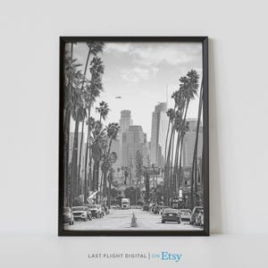 Puede incluir: Fotografía en blanco y negro de una calle de la ciudad bordeada de palmeras. El horizonte de la ciudad es visible en el fondo. Los coches conducen por la calle.