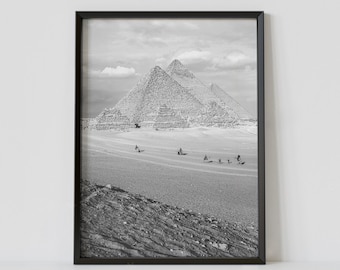 Könnte beinhalten: Ein Schwarzweißfoto der drei großen Pyramiden von Gizeh in Ägypten. Die Pyramiden sind in der Ferne, im Vordergrund eine Gruppe von Menschen, die auf Kamelen reiten.
