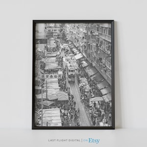 May include: A black and white photograph of a crowded street market in Hong Kong. The street is lined with stalls selling various goods, and there are many people walking around. A tram is visible in the middle of the street.