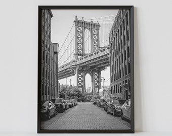 May include: Black and white photograph of a street in New York City with the Manhattan Bridge in the background. Cars are parked on both sides of the street, and the street is paved with cobblestones.