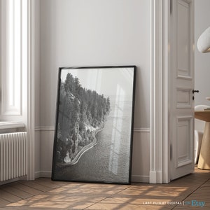 May include: Black and white aerial photograph of a winding road along a coastline with trees and a body of water.