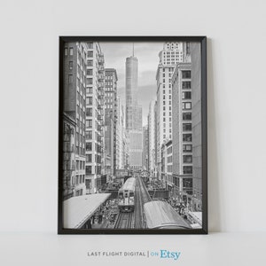 May include: A black and white photograph of a train traveling through a city street lined with tall buildings. The train is in focus, while the buildings are blurred in the background.