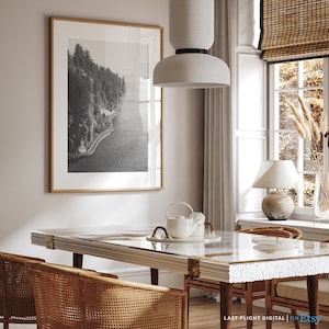 May include: A dining room with a white table and chairs, a black and white framed photograph of a coastline, a white pendant light, and a white lamp on a table next to a window.