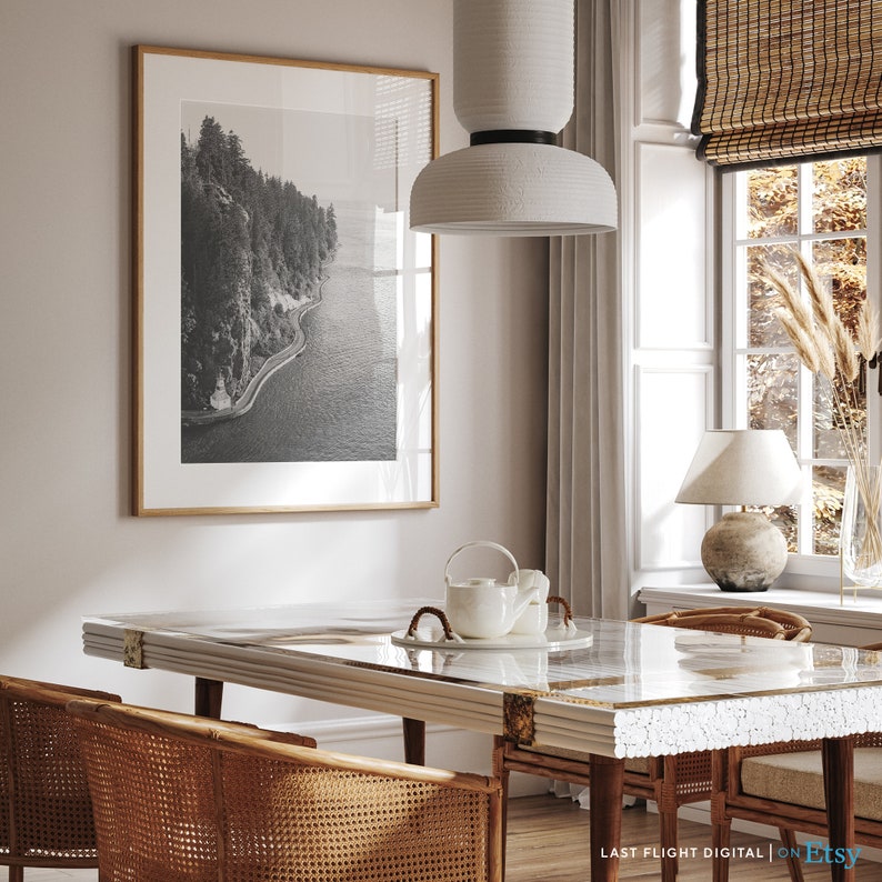 May include: A dining room with a white table and chairs, a black and white framed photograph of a coastline, a white pendant light, and a white lamp on a table next to a window.