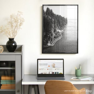 May include: A black and white photograph of a winding road along a cliffside overlooking a body of water. The photo is framed in a black frame and hanging on a white wall. A laptop computer is on a desk in front of the photo, displaying the Etsy website.