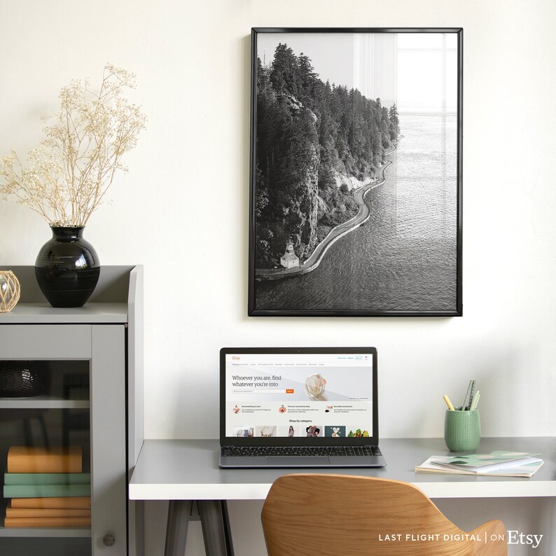 May include: A black and white photograph of a winding road along a cliffside overlooking a body of water. The photo is framed in a black frame and hanging on a white wall. A laptop computer is on a desk in front of the photo, displaying the Etsy website.
