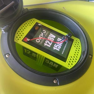 May include: A lime green battery pack with the text "12.8V 15A" and "5AH" is installed inside a black and green compartment. The battery is connected with red and black wires. The compartment is set in a yellow surface.