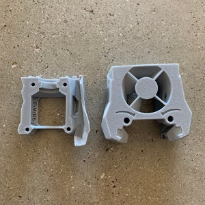 May include: Two 3D printed gray plastic parts. The part on the left has a rectangular opening and the text "E-V/R6" printed on the side. The part on the right has a circular opening with a cross-shaped design inside.