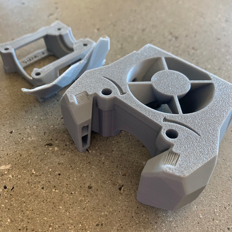 May include: A gray 3D printed fan shroud with a circular opening for a fan. The shroud has a textured surface and is designed to fit a small fan.