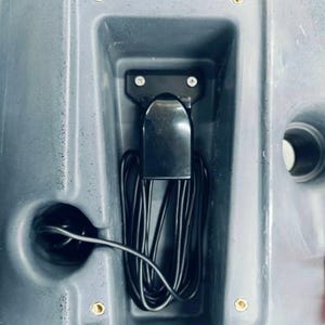May include: A close-up of a black electrical cord and a black plastic component with two screws in a gray plastic compartment.