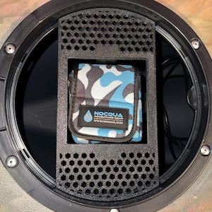 May include: A black, circular compartment with a rectangular, perforated insert. Inside, a small pouch with a blue, white, and black camouflage pattern is visible. The pouch has the text "NOCQUA ADVENTURE GEAR" and a website address.