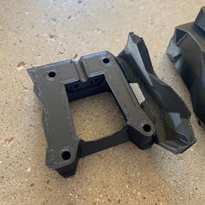 May include: Black plastic 3D printed part with a square opening and several holes. The part has a textured surface and appears to be a component for a larger assembly.