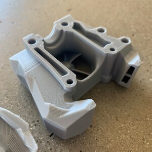 May include: A gray 3D printed plastic part with a complex design. The part has a rectangular opening and a series of ridges and grooves.