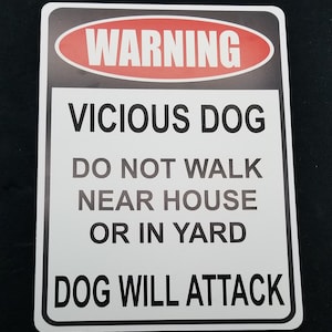 Op de afbeelding: Een wit bord met zwarte tekst en een rood ovaal waarschuwingsbord met "WARNING" in witte tekst. Het bord luidt "VICIOUS DOG DO NOT WALK NEAR HOUSE OR IN YARD DOG WILL ATTACK."