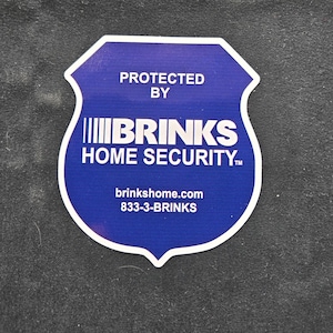 以下が含まれることがあります： 「PROTECTED BY BRINKS HOME SECURITY」の文字が入った、青と白のシールド型ステッカー。ステッカーには、ウェブサイトbrinkshome.comと電話番号も含まれています。