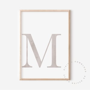 May include: A framed print featuring a large, stylized letter "M" in a muted taupe color against a white background. The print is enclosed in a light-colored wooden frame, creating a minimalist aesthetic.