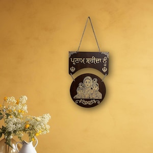 May include: A brown wooden sign with a gold inscription in Punjabi that reads "Puttam Sarita Hai" and a gold image of a family with three people. The sign is hanging on a yellow wall.