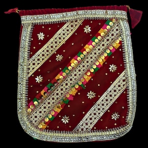 Puede incluir: Una bolsa de terciopelo rojo con adornos dorados y plateados. La bolsa está decorada con un patrón diagonal de ribetes dorados y plateados, lentejuelas y borlas de colores.
