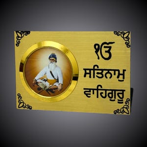 Puede incluir: Una placa de color dorado con una imagen circular de un gurú sij con turbante y barba. La placa tiene el texto "ੴ ਸਤਿਗੁਰ ਪ੍ਰਸਾਦਿ" en letras negras.