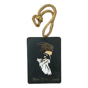 May include: Black car air freshener with a gold image of a man with a beard and the text "Mera Baba Nanak". The air freshener is attached to a gold string.