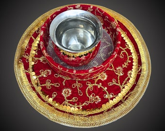 May include: A red velvet thali with gold trim and sequins, featuring a small silver bowl in the center. The thali is used for traditional Indian ceremonies.