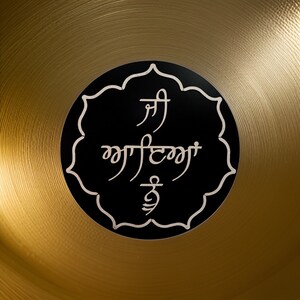 Può includere: Una ciotola cantate dorata con un adesivo nero sul fondo. L'adesivo presenta un design bianco con le parole "Sri" e "Waheguru" in scrittura Gurmukhi.